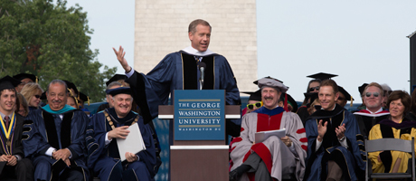 Brian Williams speaking a podium at GW commencement with the Washington Monument behind him