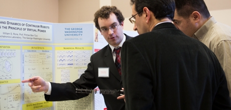 student points to presentation board at showcase with two people reviewing his work