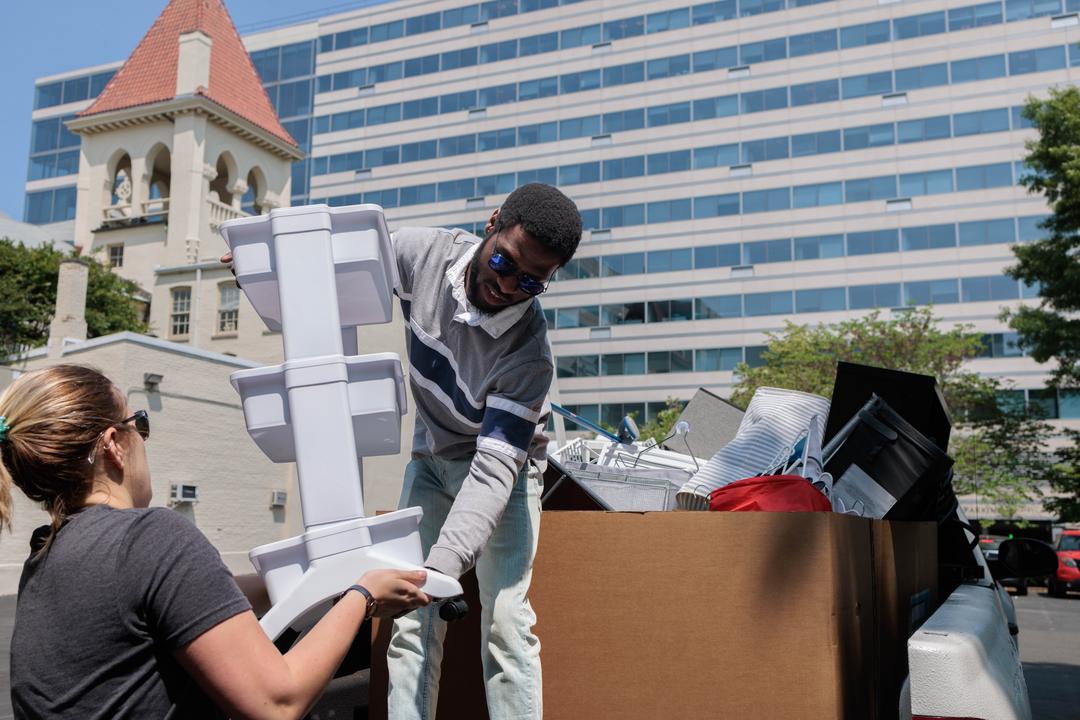 A white woman helps a Black man move a plastic shelf from the bed of a truck to a storage closet. 
