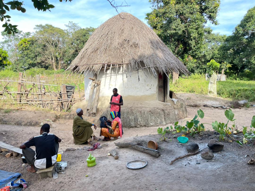 A Clinic+O health care worker in a pink vest talking to 3 people in traditional clothing who are sitting on the ground in a Guinea village. A grass hut and a field & trees are behind them,.