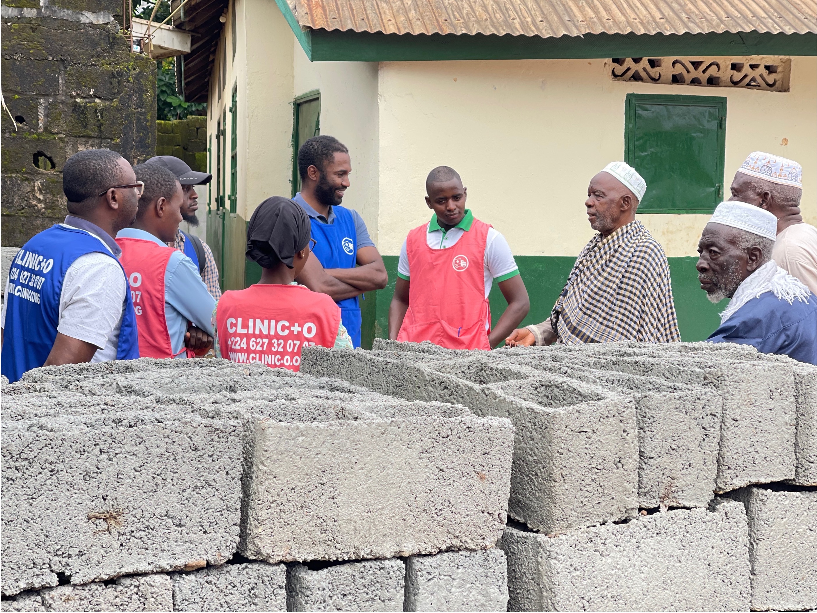 Nasser Diallo in blue vest surrounded by several other men in blue & pink vests & traditional clothing in an Guinea village with a house behind them & cinder blocks in front of them.