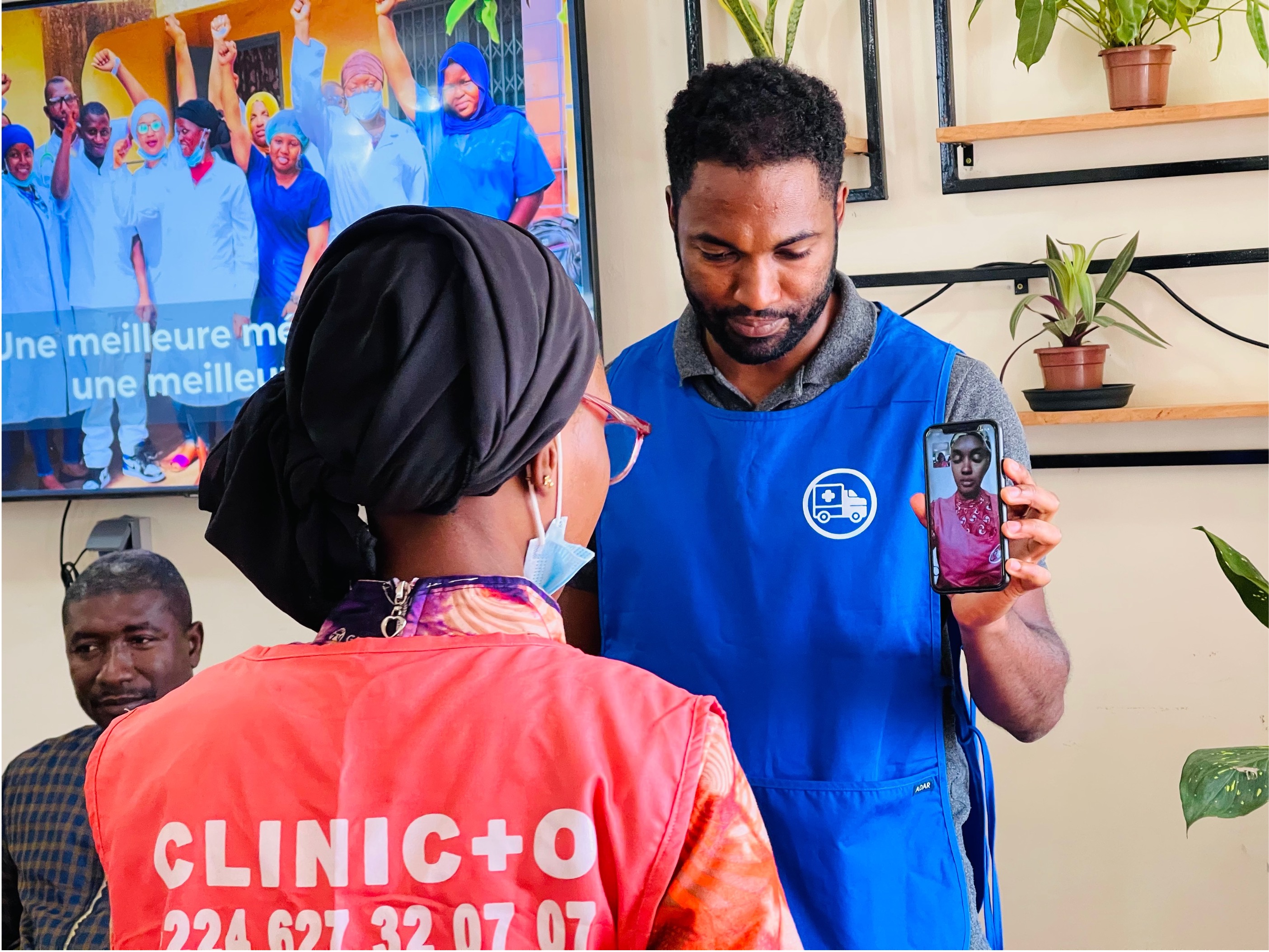 Nasser Diallo in blue vest showing a Clinic+O health worker in a pink vest how the mobile app works. He's holding a phone in one hand in his clinic.