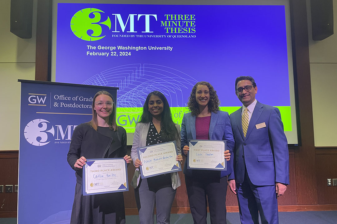 3 female students and a man in a suit in front of the blue & green 3MT background, (from left) Caitlin Bailey, Prachi Mahableshwarkar and Leah Kaplan with Vice Provost for Graduate and Postdoctoral Affairs Suresh Subramaniam. 