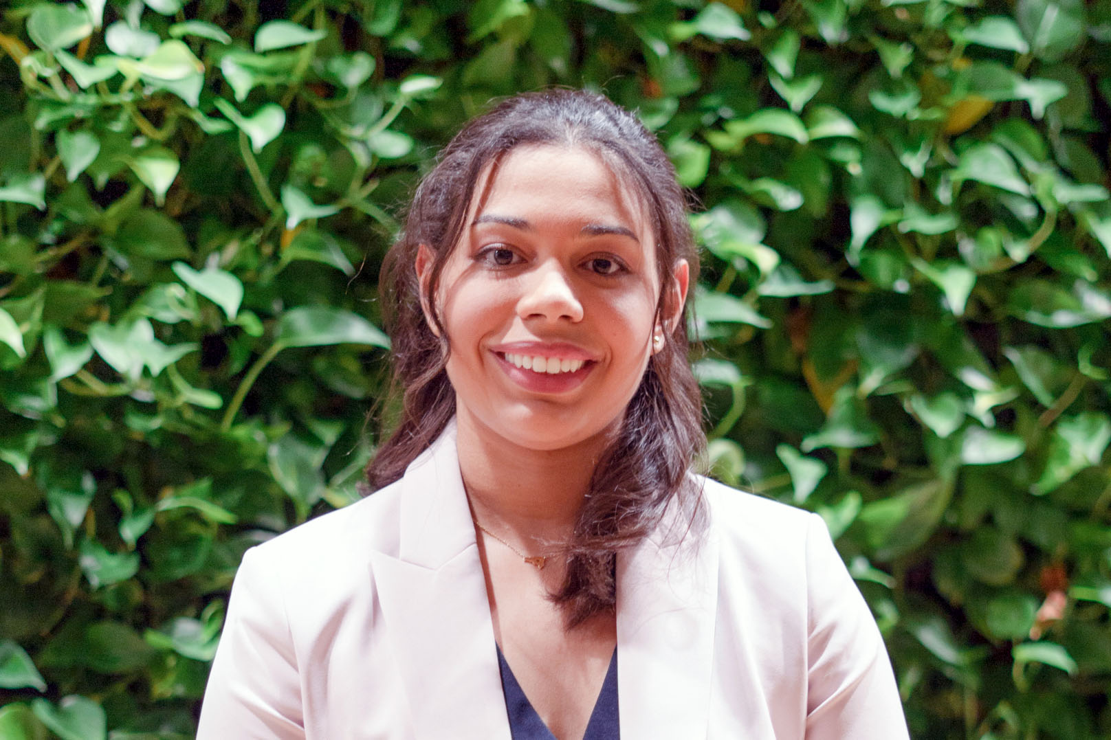 Claudia Melo. Dark hair, white jacket, blue blouse in front of leaf wall at SEH
