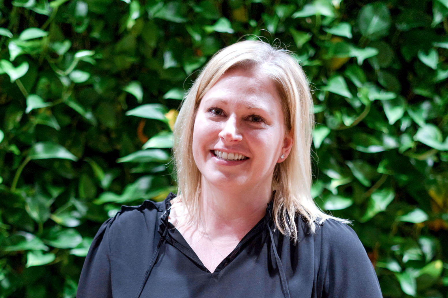 Heather Walsh. White woman blonde hair in black blouse in front of leaf wall at SEH