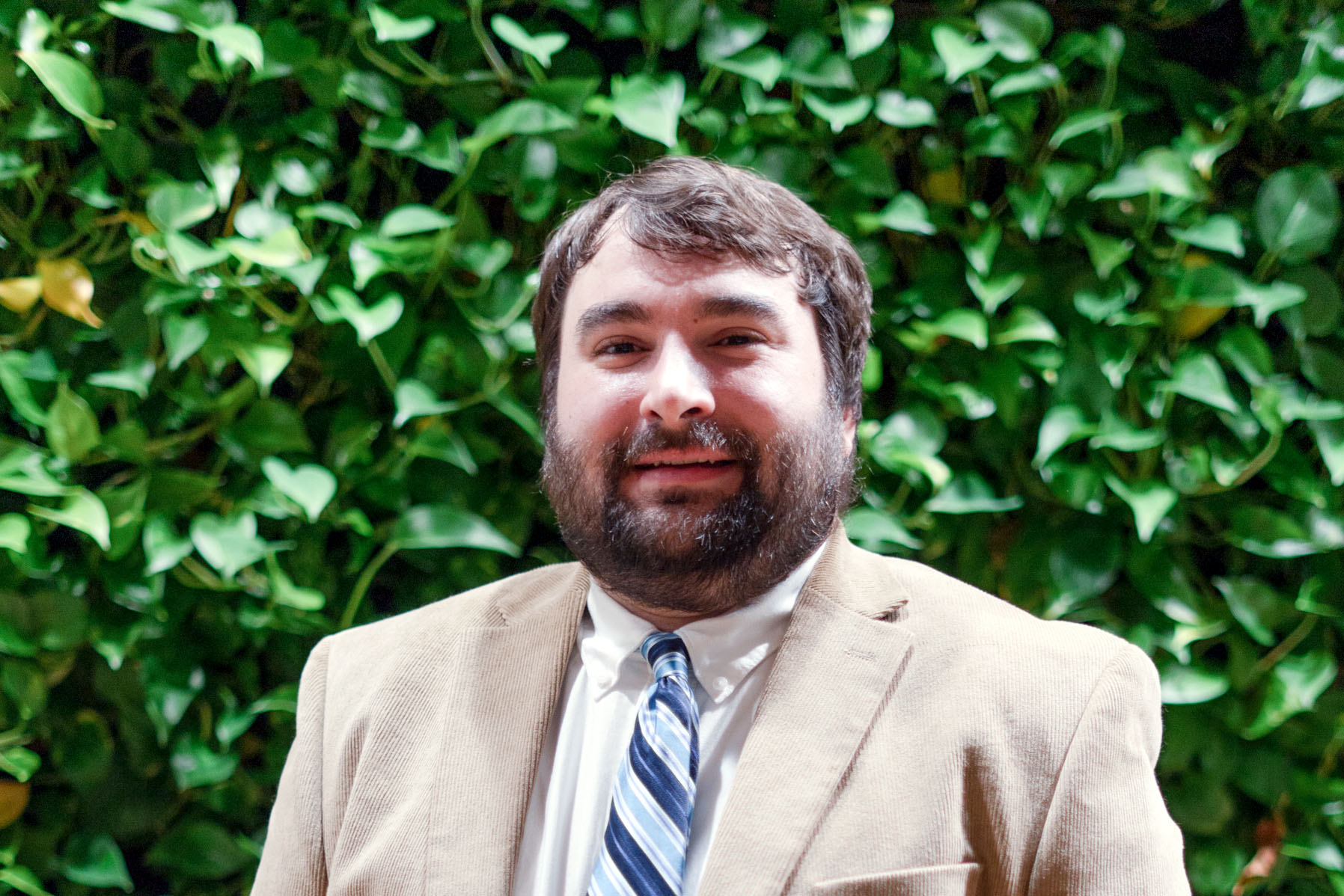 Justus Jobe. White man with bear in tan jacket & blue striped tie in front of lead wall at SEH