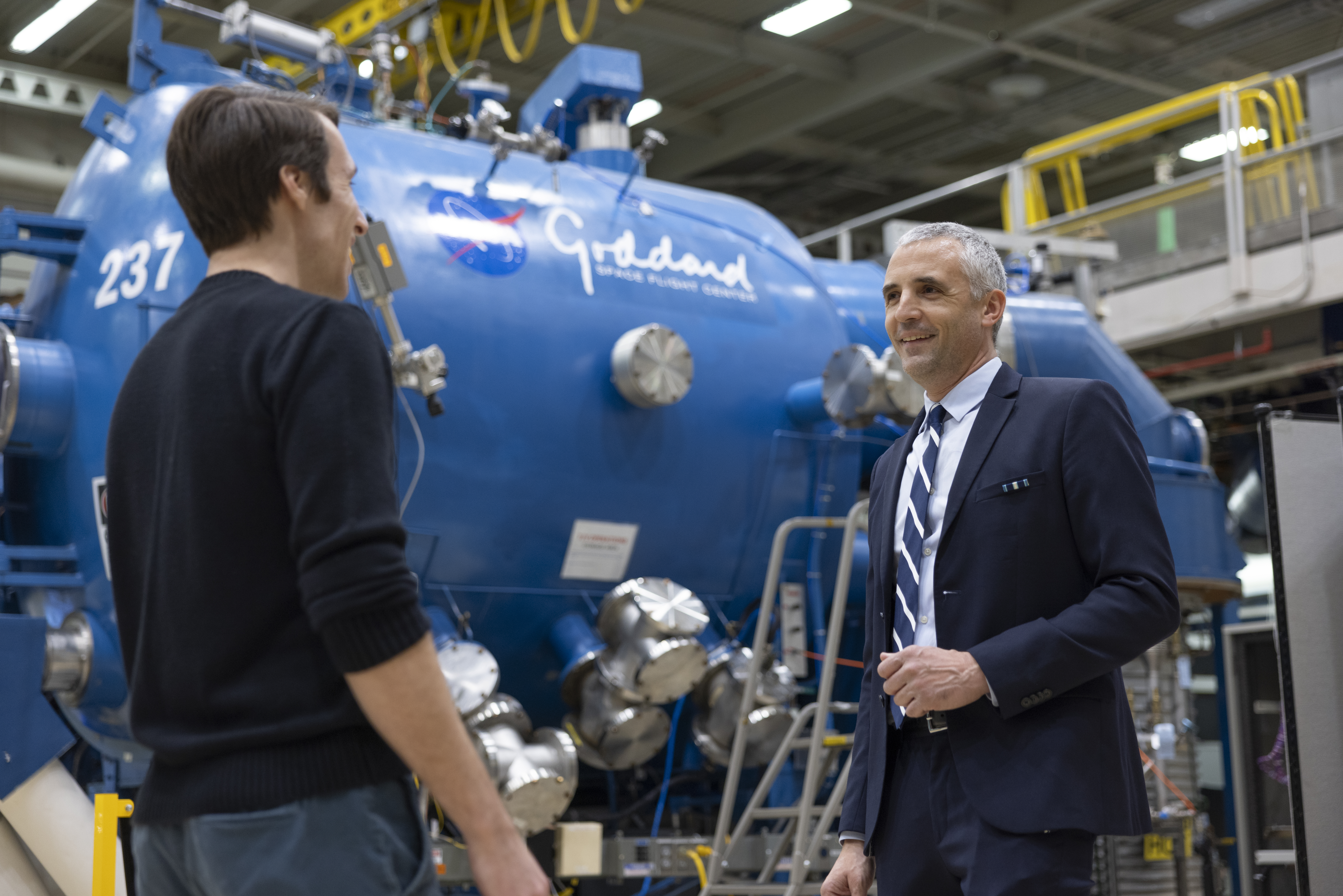 Nick Kirschner in a blue shirt facing Sylvain Guiriec in a blue suit at NASA’s Goddard Space Flight Center in front of a blue tank