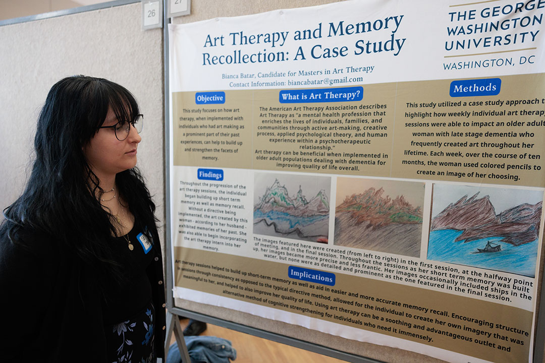 Art therapy student Bianca Bater in a black dress with black hair & glasses in front of her poster that shows drawings of mountains in Japan.