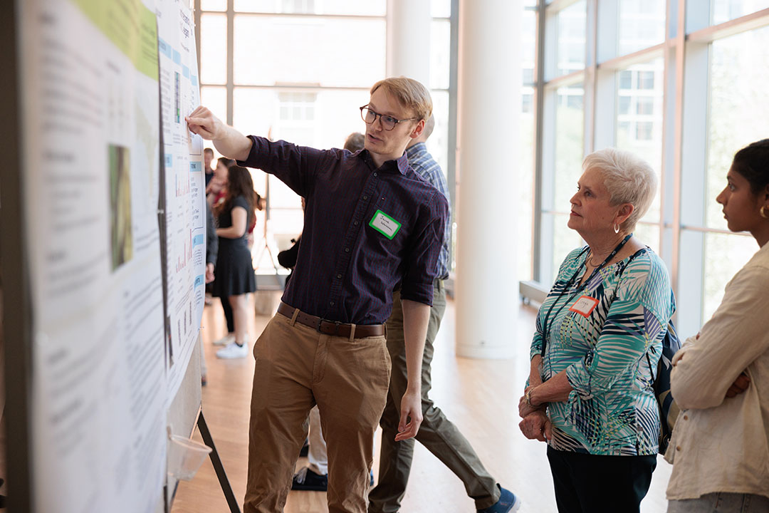 Junior biology major Jacob Washton in a blue shirt and tan slacks shows a poster project on a cork board to President’s Spouse Sonya Rankin in a blue flowered blouse and black pants.
