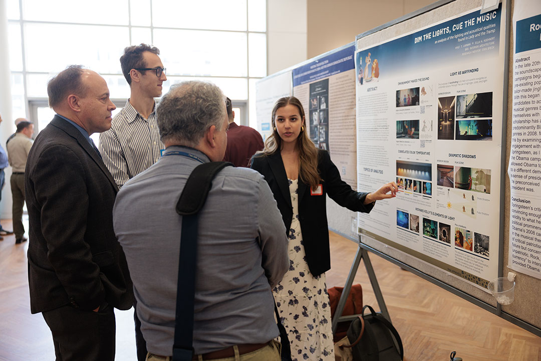 4 people in front of a cork board with a student poster at the university student center, including (from left) CCAS Dean Paul Wahlbeck in a dark suit, student Kyle Layman (tall man with classes) & student Ella Kuehnert ( in white dress with blue blazer) & a grey haired man with his back to the camera in a blue dress shirt.