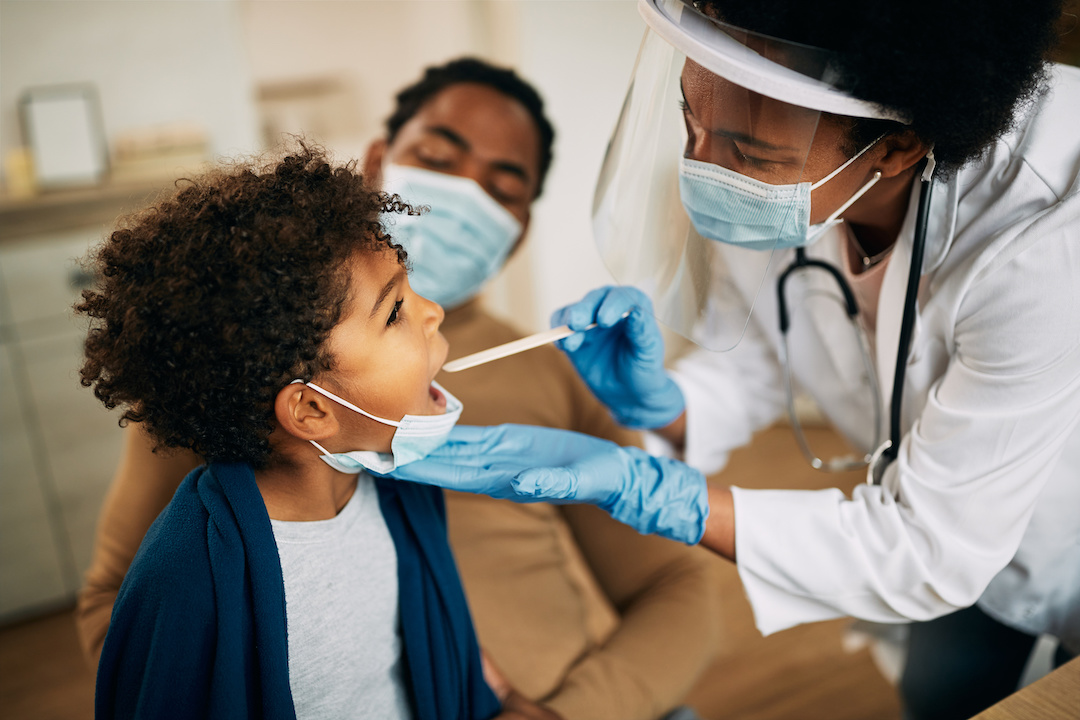 A doctor examines a child patient with a tongue depressor while the child's parent looks on. (Adobe)