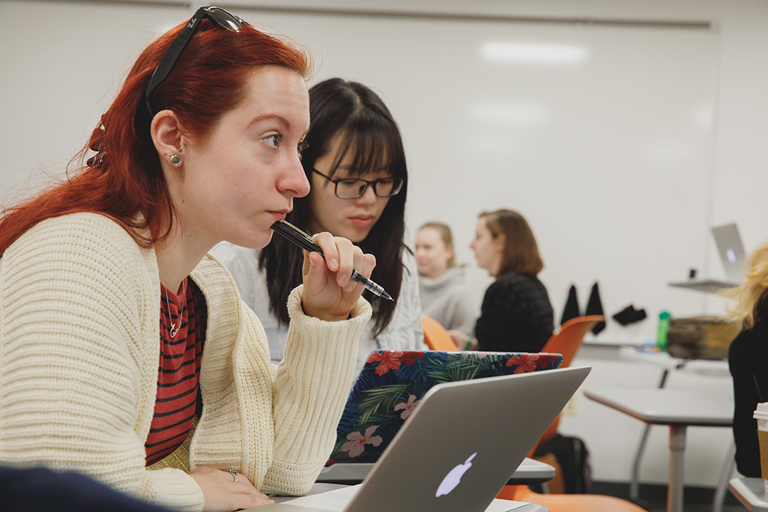 Two female students in a classroom with an open laptop: Red hair, white sweater. Black hair & black glasses