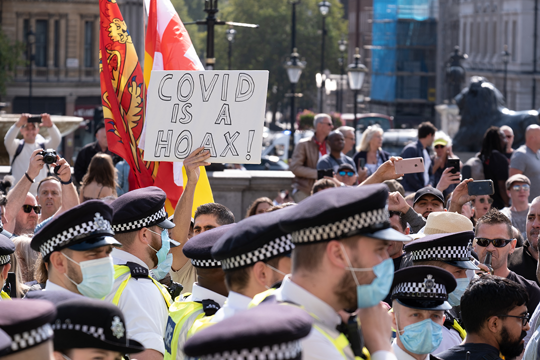 2020 Trafalgar Square protest against COVID-19 restrictions. Includes a crowd, the heads of several police and someone holding up a white sign that reads "COVID IS A HOAX."