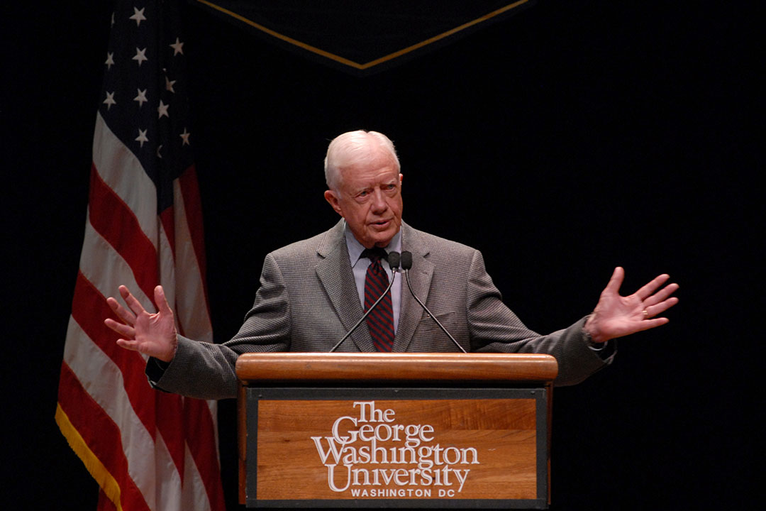 A photo of Jimmy Carter in a grey suit at a GWU lectern with an American flag behind him