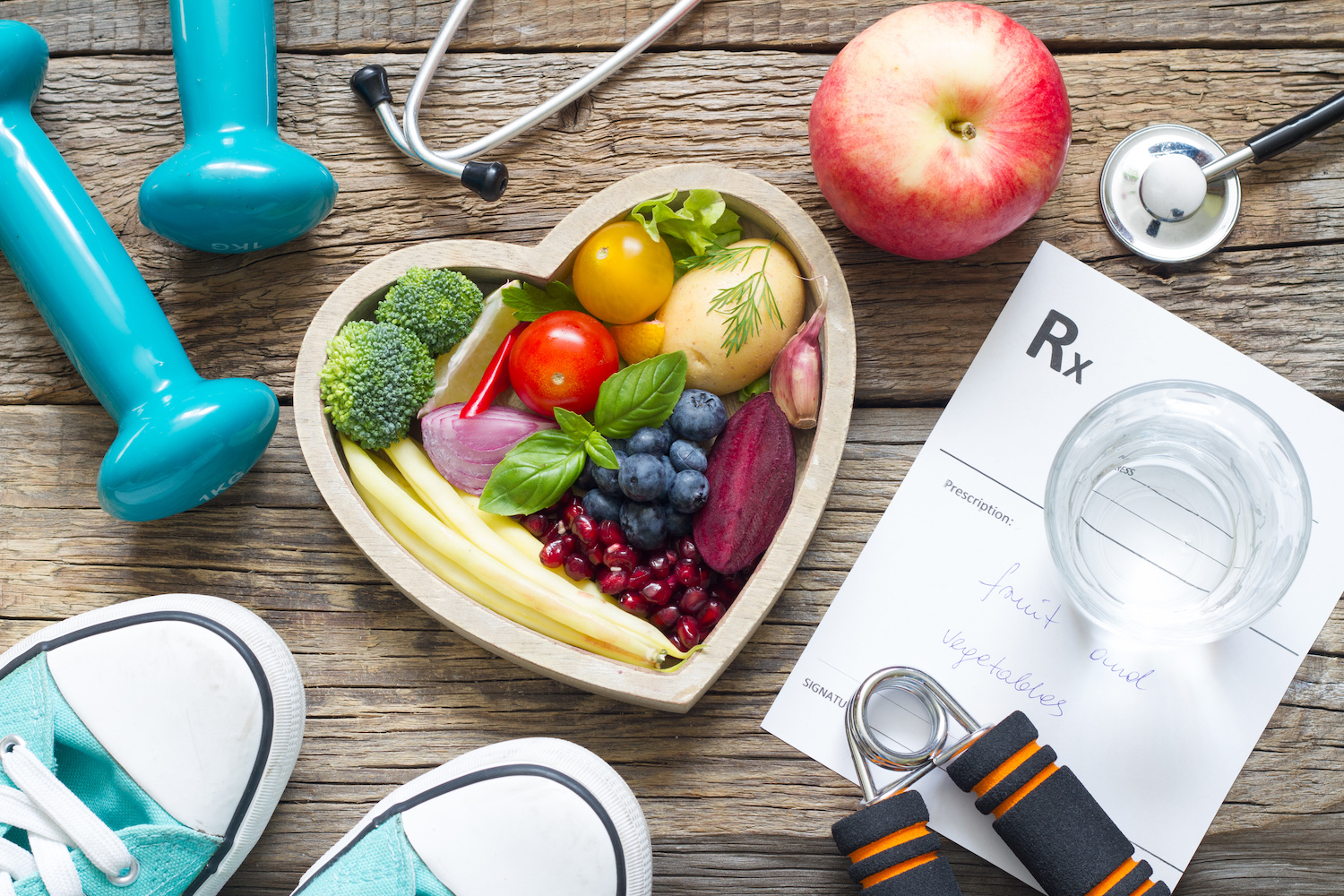 Heart-healthy foods arranged in a heart shaped bowl, with weights, a stethoscope, workout shoes and glass of water. 