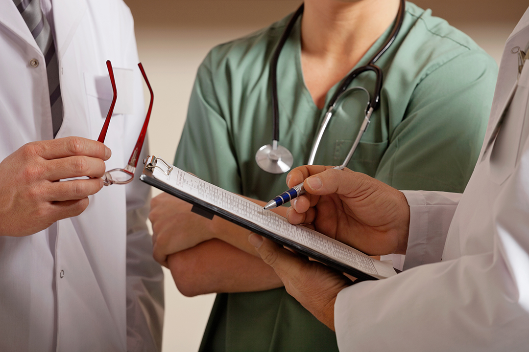 Three people in scrubs consult a medical chart. Their faces are not visible (Adobe Stock)
