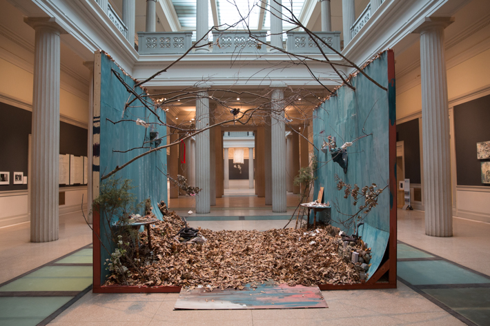 A whimsical installation featuring two turquoise walls adorned with overhanging branches, antlers, candles and art. A pile of dried leaves covers the floor.