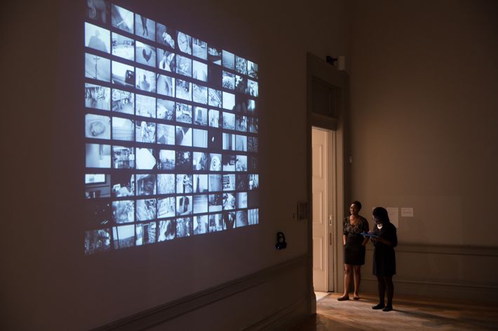 Two guests stand in the lit doorway of a darkened room. The wall adjacent projects a grid of screens, Kesi Marcus' short film installation.