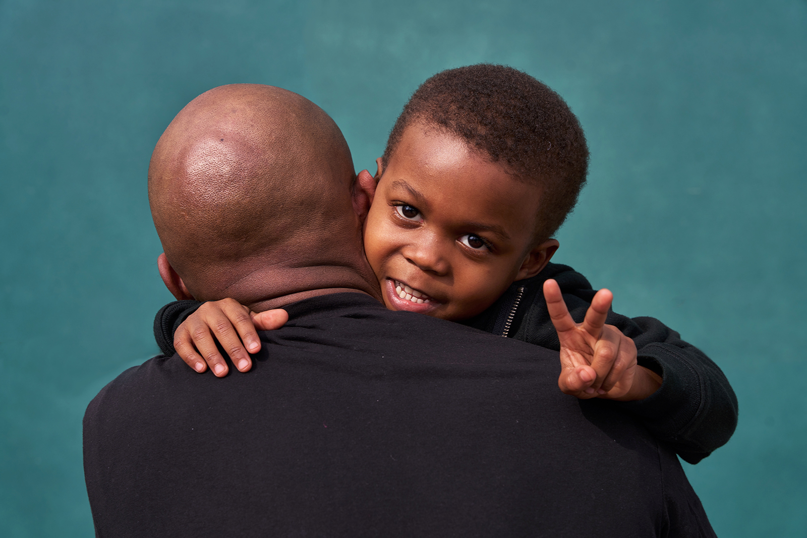 Color photo, a child flashes a peace sign over a man's shoulder