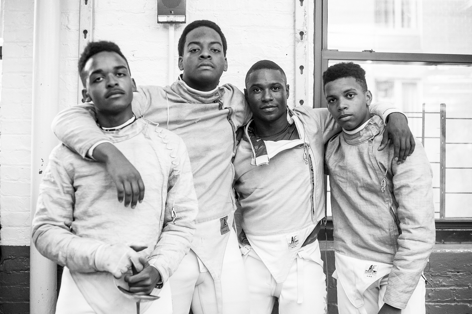 Four young black men in fencing gear with their arms around each others' shoulders look into the camera