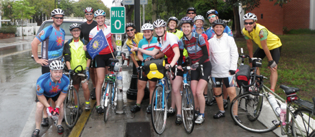 group of bicycle riders crowds around a Mile 0 sign on a street smiling 