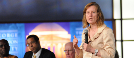 Janis Orlowski speaks with microphone in hand with two men seated in background 