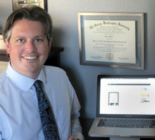 Derek Walter in office standing next to laptop and framed degree from the George Washington University