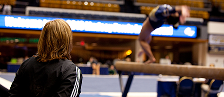 Member of gymnastics team does a back flip on balance beam with coach looking on from sidelines