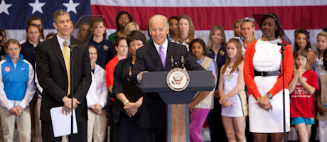 Joe Biden at podium speaking to audience with Arne Duncan and students of a variety of ages looking on 