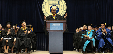 Adm. Mel Williams speaks at podium in front of university members on stage in regalia
