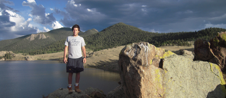 male wearing George Washington University shirt stands in mountains overlooking body of water