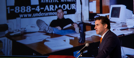 Kevin Plank speaks at podium with a background of a pic of him from the early days of Under Armour