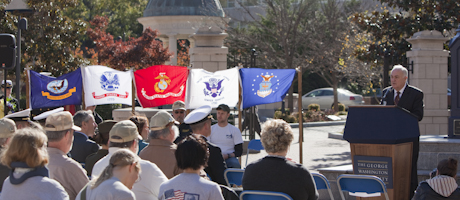 Steven Knapp speaks at podium in Kogan Plaza on veterans day with military flags & members of the university community watching