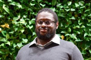 Michael Guy. Black man with glasses & beard & grey sweater in front of leaf wall at SEH