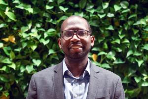 Travis Reginal. Black man with glasses in grey jacket & blue in front of leaf wall at SEH
