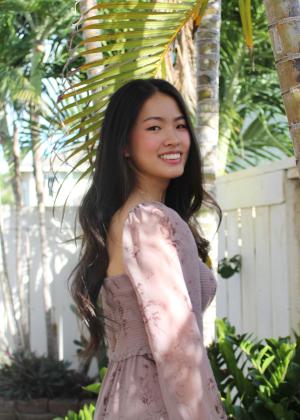 CCAS philosophy major Seyeon Moon in a pink dress and long black hair smiling standing in front of a tree and fence.
