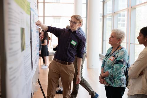 Junior biology major Jacob Washton in a blue shirt and tan slacks shows a poster project on a cork board to President’s Spouse Sonya Rankin in a blue flowered blouse and black pants.