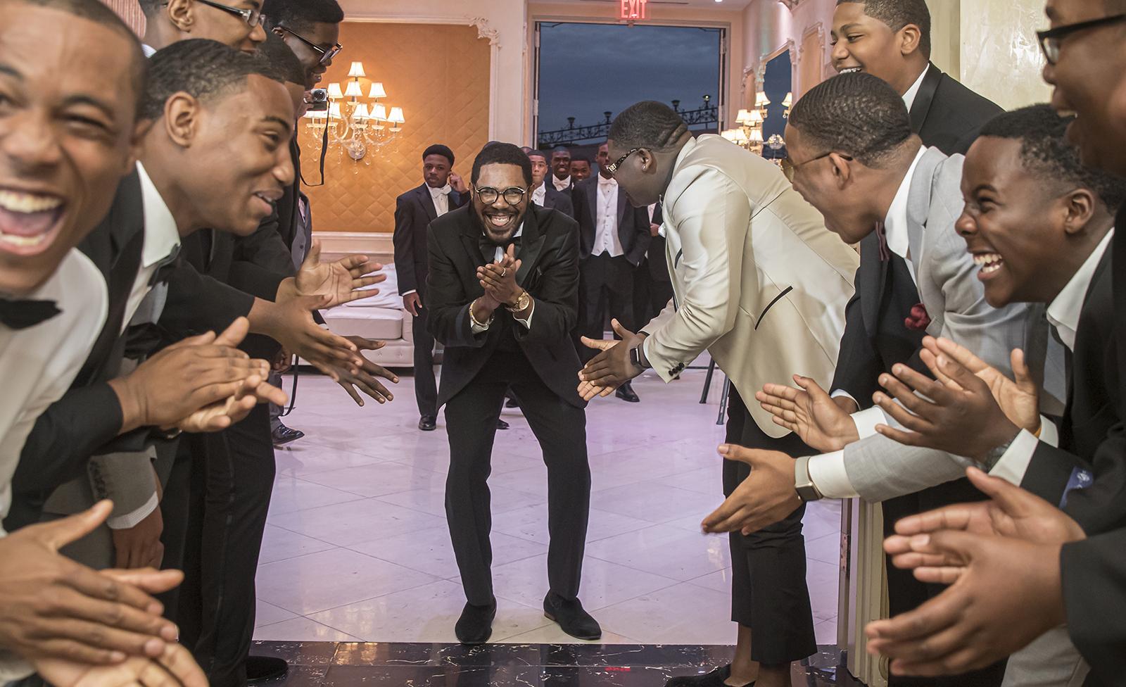Group of Black men in suits applauding and laughing in two lines, framing a single man