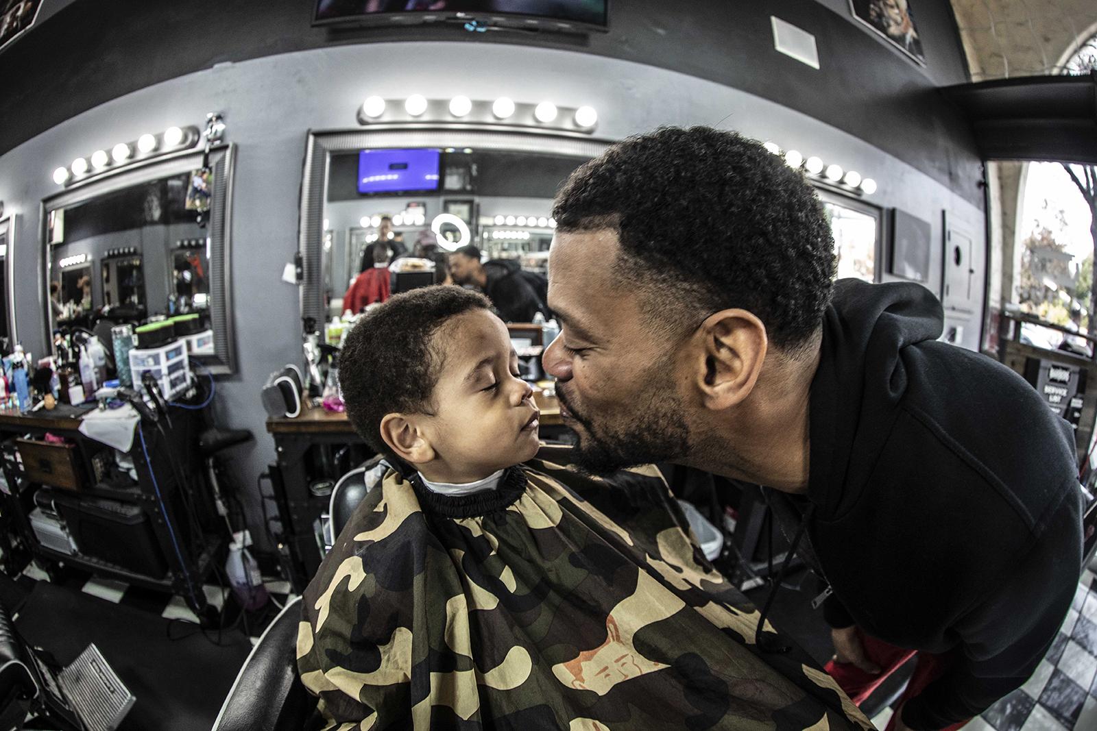 Color photo, fisheye lens image of a smiling father kissing his son who sits in a barber's chair