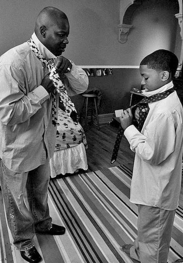 Black and white photo, a man demonstrates how to tie a necktie to a boy