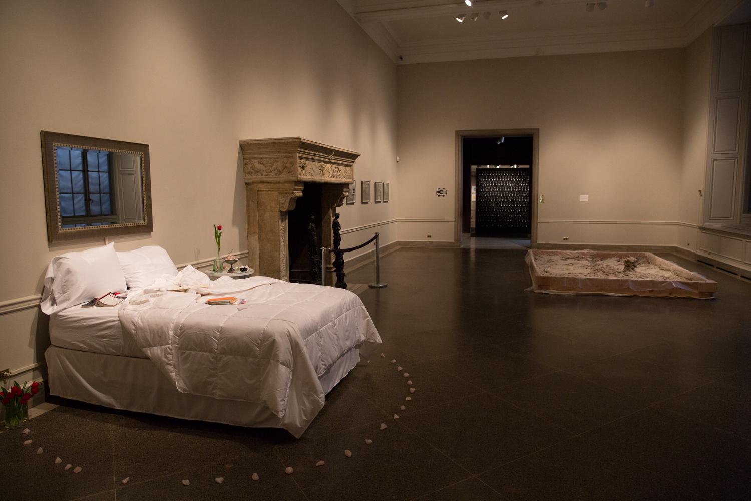 A bed, encircled by stones, sits against a wall with a fireplace to its left.