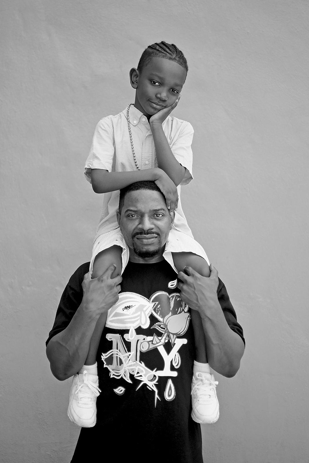 Black and white photo, a man holds a child on his shoulders, both are gazing at the camera