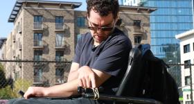A man with dark curly hair and glasses sits on a wheelchair and presses a button on a camera rubber-banded to the wheelchair's armrest.