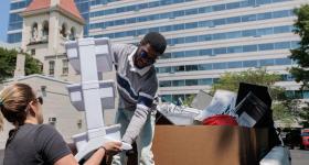 A white woman helps a Black man move a plastic shelf from the bed of a truck to a storage closet. 