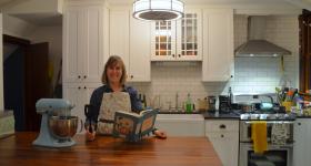 Caroline Smith in her kitchen. Standing at her table with a mixer next to her and a book open.