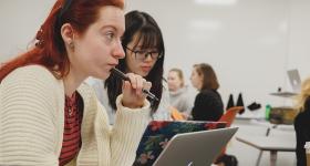 Two female students in a classroom with an open laptop: Red hair, white sweater. Black hair & black glasses