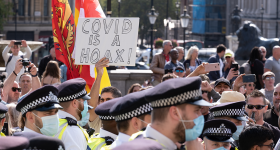 2020 Trafalgar Square protest against COVID-19 restrictions. Includes a crowd, the heads of several police and someone holding up a white sign that reads "COVID IS A HOAX."