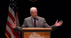 A photo of Jimmy Carter in a grey suit at a GWU lectern with an American flag behind him
