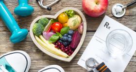 Heart-healthy foods arranged in a heart shaped bowl, with weights, a stethoscope, workout shoes and glass of water. 